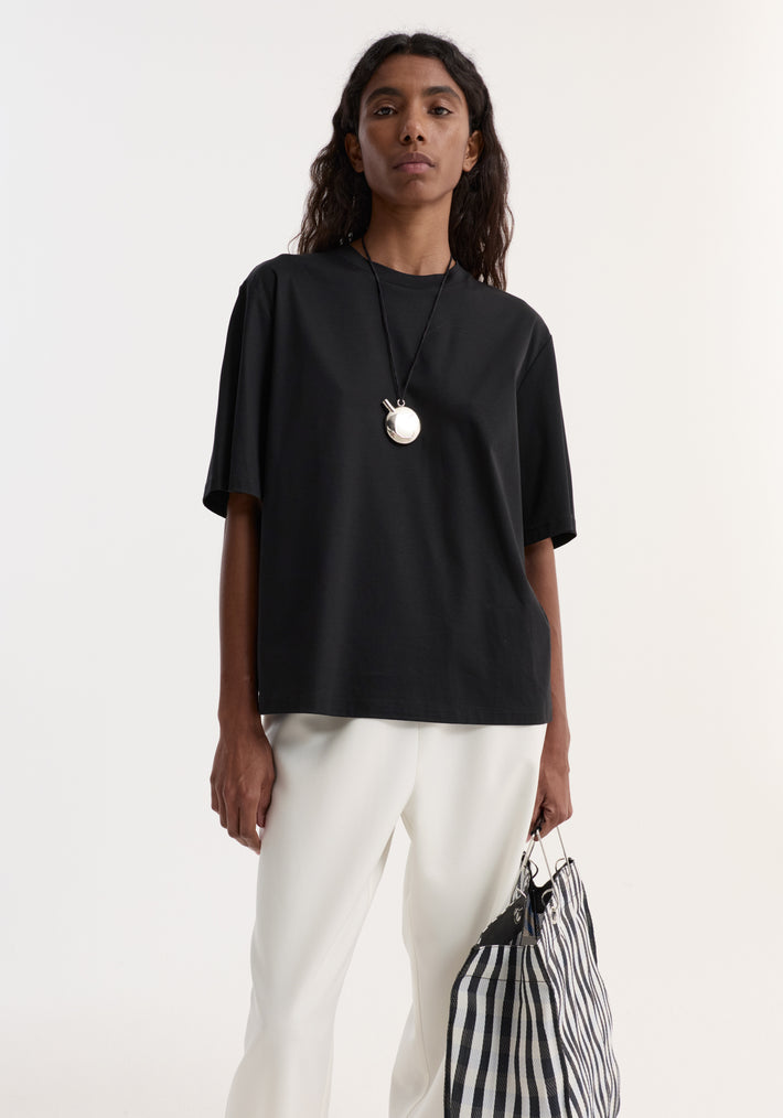 Person wearing an Oversized cotton t-shirt | black with a loose fit and drop shoulder, paired with white trousers, holding a striped tote bag.