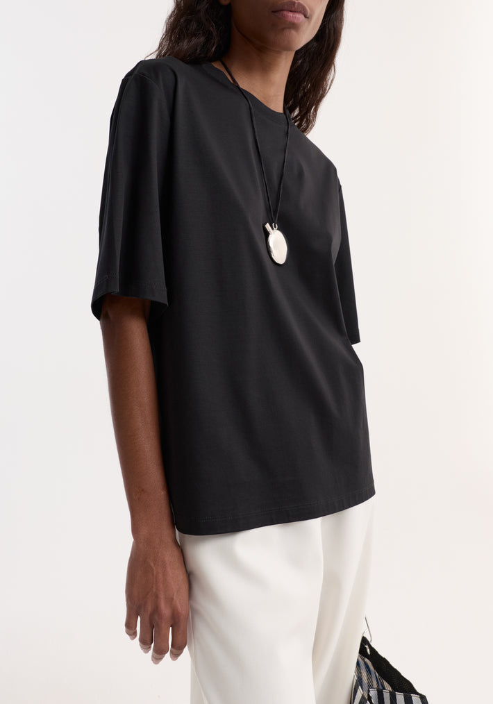 Person wearing an Oversized cotton t-shirt | black, featuring an oversized fit, drop shoulder, and crew neck, tucked into light-colored trousers with an oval pendant necklace.