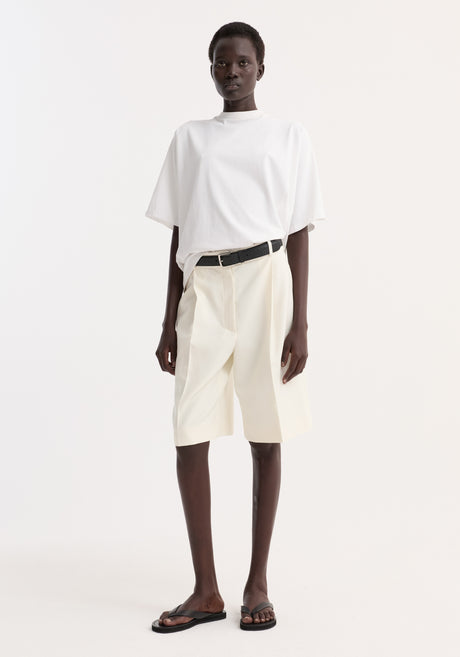 Model wearing Oversized cotton t-shirt in optic white, tucked into cream shorts, with a black belt and sandals. Features relaxed fit, drop shoulder, crew neck.