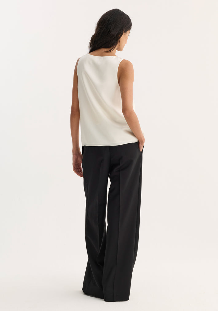 Woman in scoop-neck silk top, cream, from behind, showcasing loose fit and sleeveless design, paired with wide-leg trousers, hand in pocket, hair flowing.