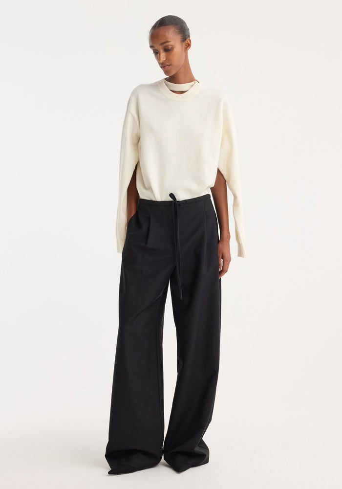 Model wearing Relaxed drawstring trousers in black, featuring a wide-leg silhouette, drawstring waistband, front pleats, and back welt pockets, paired with an ivory sweater.