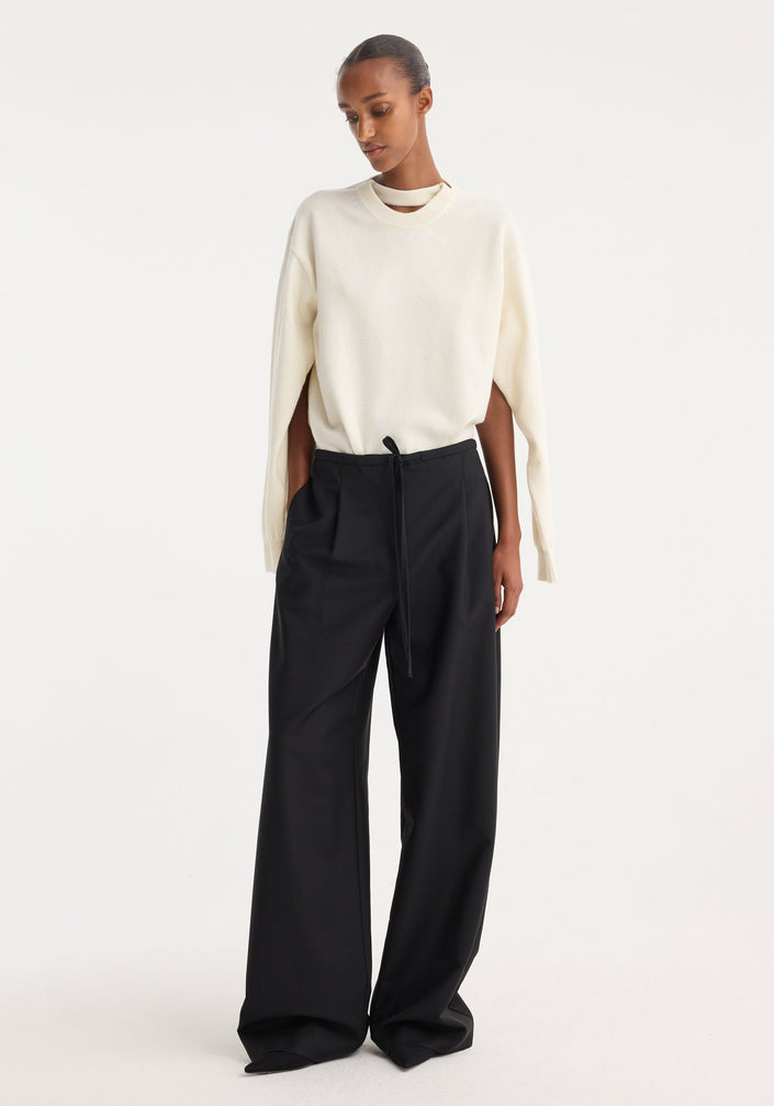 Model wearing Relaxed drawstring trousers in black, featuring a wide-leg silhouette, drawstring waistband, front pleats, and back welt pockets, paired with an ivory sweater.