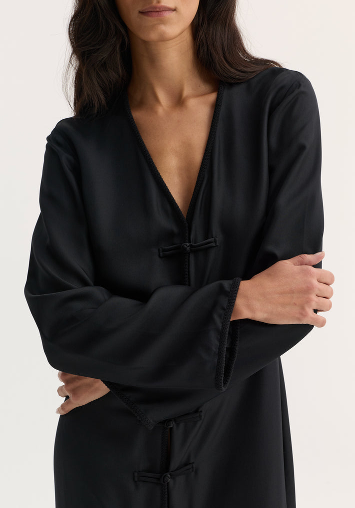 Mandarin silk dress in black, featuring a V-neckline, frog-style closures, and satin finish. Model's arms folded, showcasing elegant silk craftsmanship.