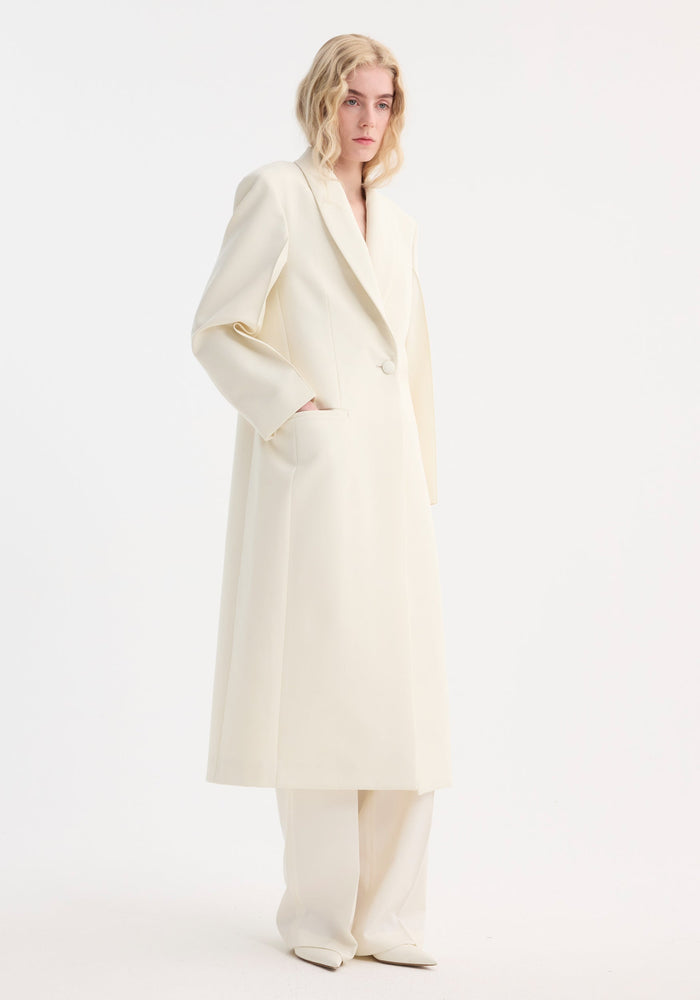Tall person in a Signature Opera Coat | Ivory, showcasing a fitted, long coat with sleek lapels, single-breasted closure, and welt pockets.