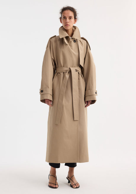 Model in Oversized classic trench | clay, featuring double-breasted design, epaulettes, belted waist, and side pockets, over black cropped trousers and strappy sandals.