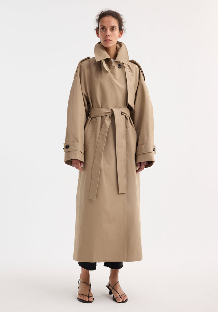 Model in Oversized classic trench | clay, featuring double-breasted design, epaulettes, belted waist, and side pockets, over black cropped trousers and strappy sandals.