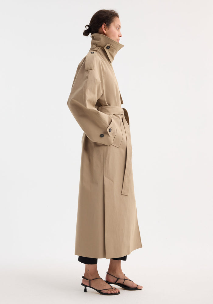 Side view of a woman in an Oversized classic trench | clay, featuring epaulets, a buttoned collar, and a cinched waist, paired with black trousers and sandals.