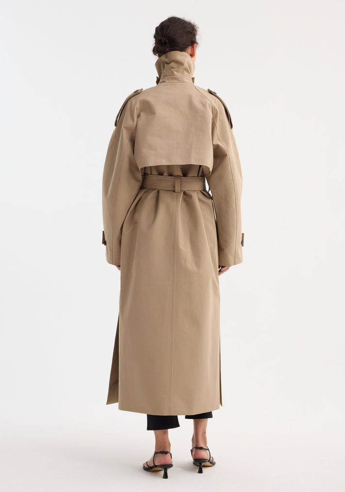 Person wearing Oversized classic trench | clay, featuring a full-length, sand-beige design with high collar, epaulettes, storm flap, and self-tie belt, paired with black trousers and sandals.