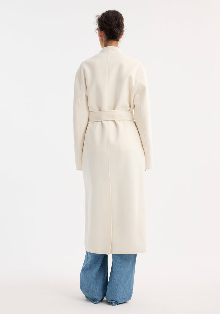 Back view of a person wearing the Double-faced kimono style coat in off white, featuring a self-tie belt and patch pockets, over light-blue jeans.