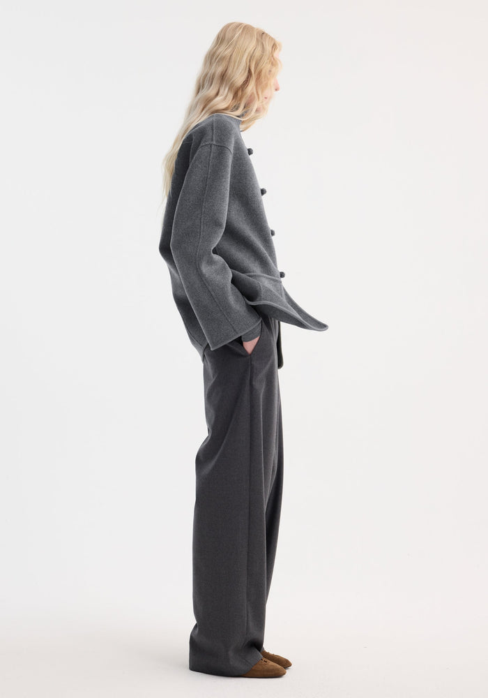 Side-profile of a model wearing the Róhe 'Marie' Mandarin double-faced wool jacket in mid grey melange, featuring toggle buttons and patch pockets, paired with matching trousers.