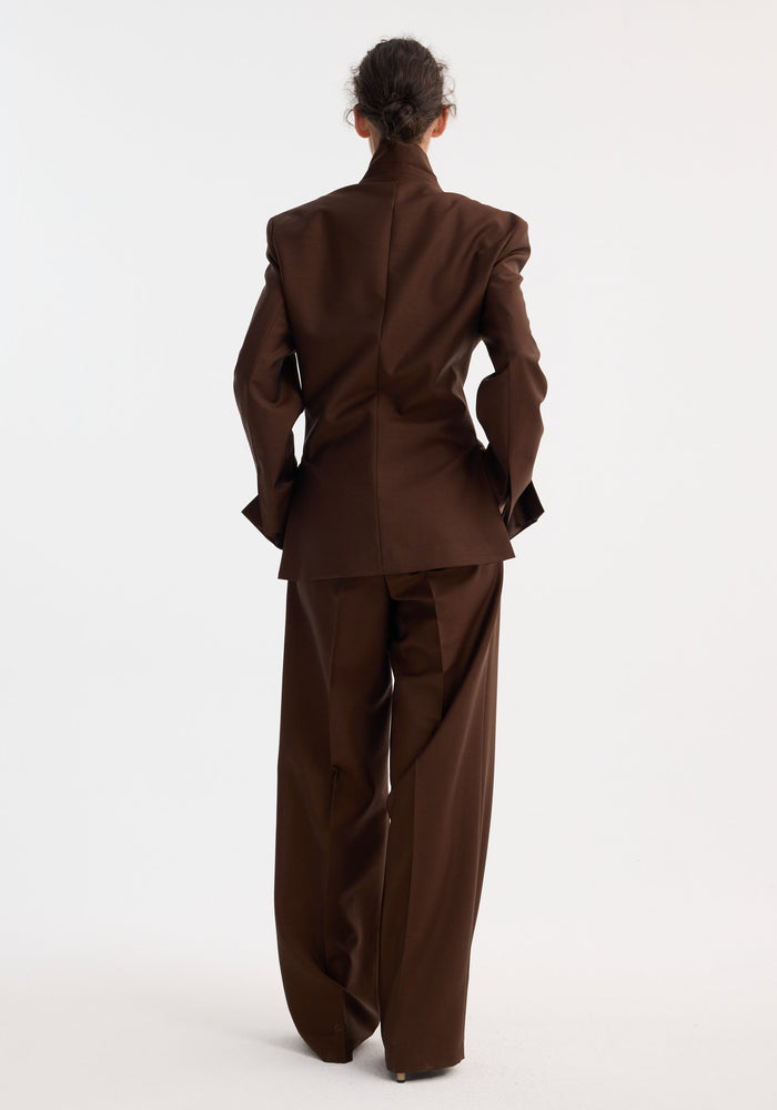 Person wearing Dual tone piqué overlap suit jacket in mahogany, featuring an oversized blazer and wide-leg trousers, standing against a plain studio background.