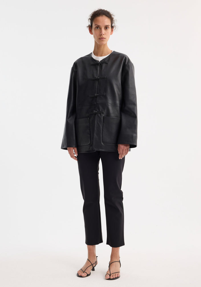 Mandarin leather jacket in black with pankou closures, patch pockets; model wears it with slim trousers and strappy sandals, showcasing the jacket's sleek design.