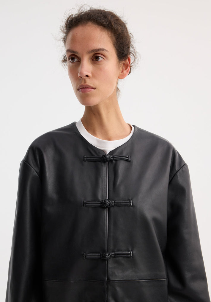 Young woman wearing the Mandarin leather jacket | black, featuring pankou closures and a plain white shirt underneath, set against a simple studio backdrop.