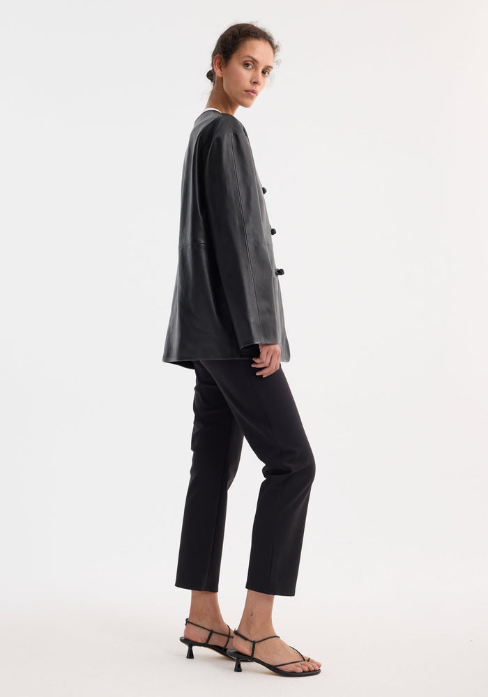 Young woman in profile wearing the Mandarin leather jacket with pankou closures, paired with slim trousers and sandals, glancing over her shoulder.