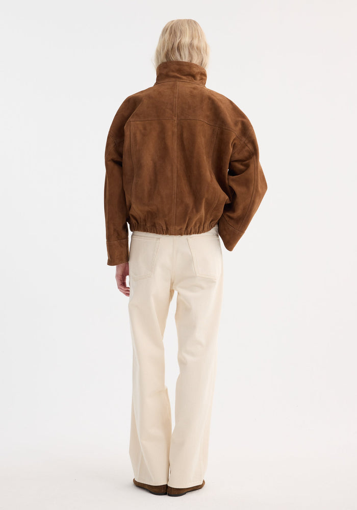 Back view of person in a Reinvented suede jacket | cognac, featuring a high collar, dropped shoulders, elasticated hem, paired with straight-leg trousers and brown shoes.