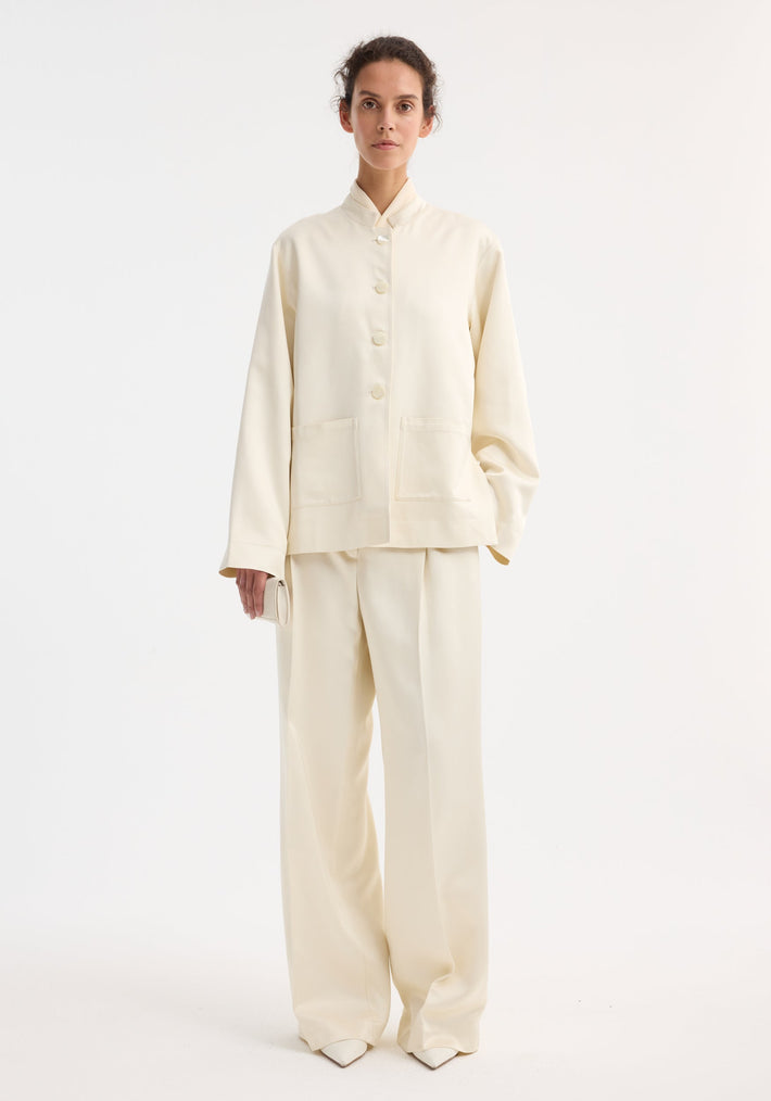 Woman in Mandarin pearl button satin jacket, cream, with high-waisted trousers and clutch. Loose fit, mandarin collar, pearl buttons, front patch pockets.