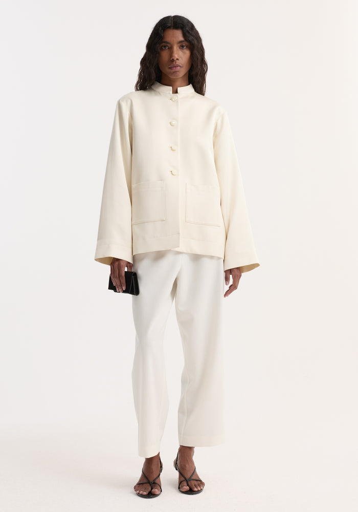 Model in Pearl button satin jacket | cream, featuring a stand collar, pearl buttons, and patch pockets, paired with matching straight-leg trousers and black sandals.