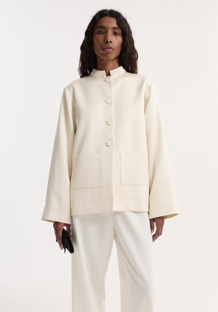 Pearl button satin jacket in ivory-cream with mandarin collar and patch pockets, worn with white trousers. Model holds a black clutch.