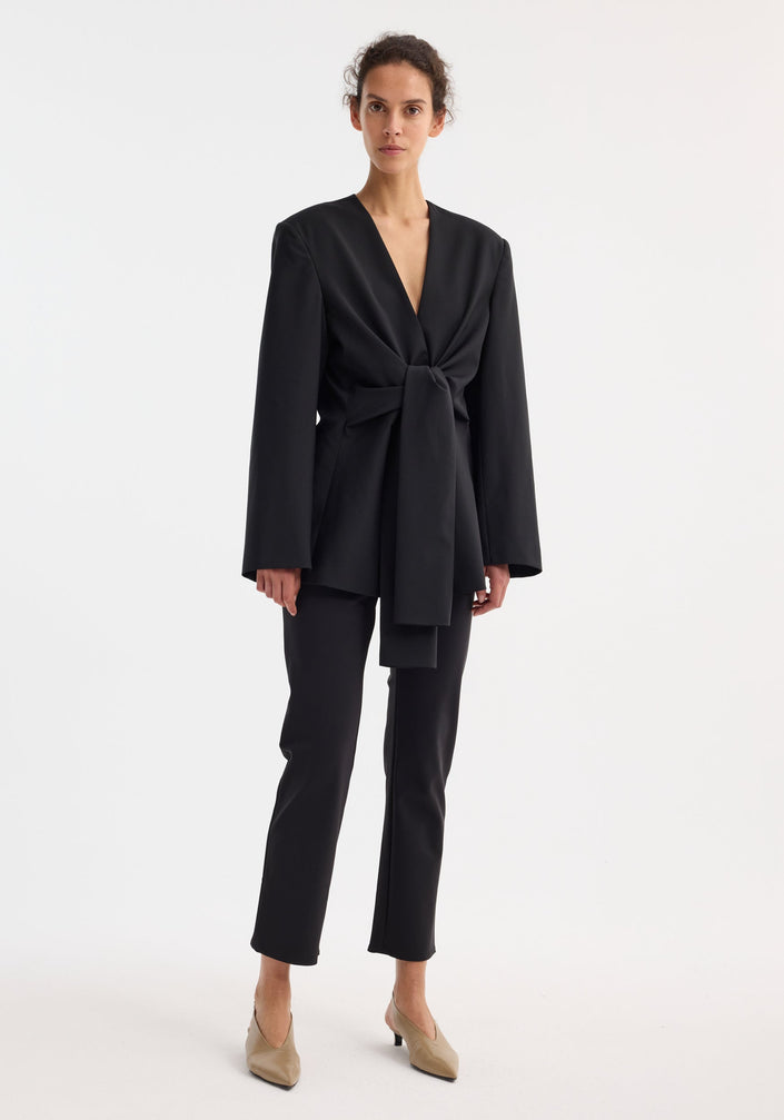 Sleeve-cinched blazer-top in black with deep V-neck and self-tie waist; combines tailoring structure with elegant wrap design. Brand label “RÓHE” visible at back neck.