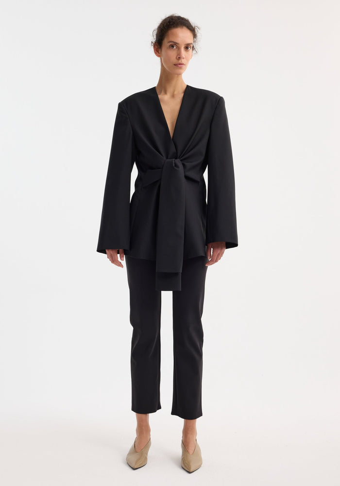 Woman wearing a Sleeve-cinched blazer-top in black, featuring a deep-V wrap design with a waist knot, paired with slim black trousers and pointed neutral shoes.