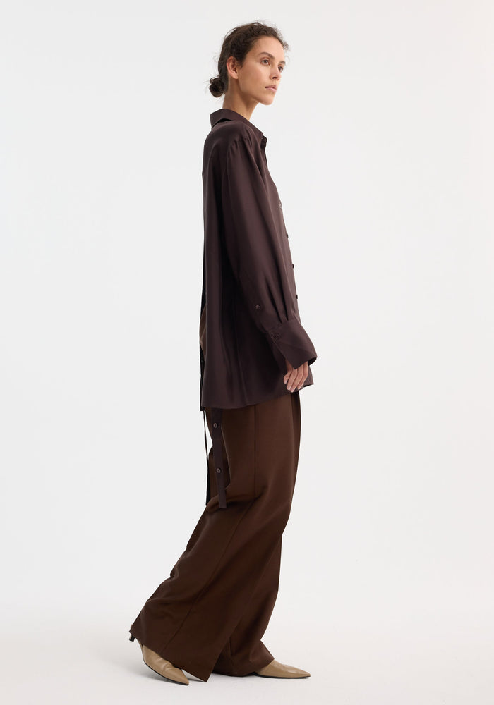 Model in profile wearing Deconstructed silk shirt | mahogany with open back and tie details, paired with matching trousers and beige ankle boots.