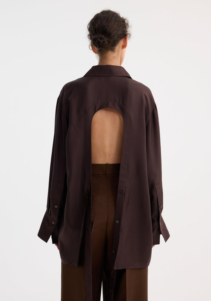 Person wearing Deconstructed silk shirt | mahogany, showcasing its open back with tie detail, paired with matching trousers. Hair in low bun, shirt drapes past hips.