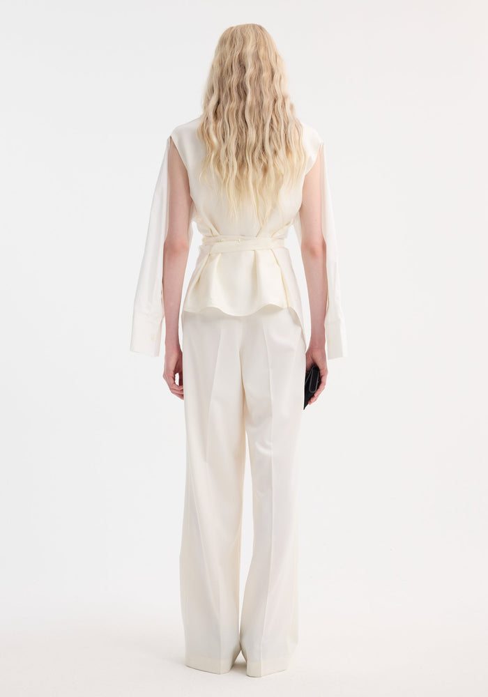 Model in back view wearing an ivory Poplin silk combined gilet-shirt with peplum flare, dramatic sleeves, and high-waisted wide-leg trousers, holding a black clutch.