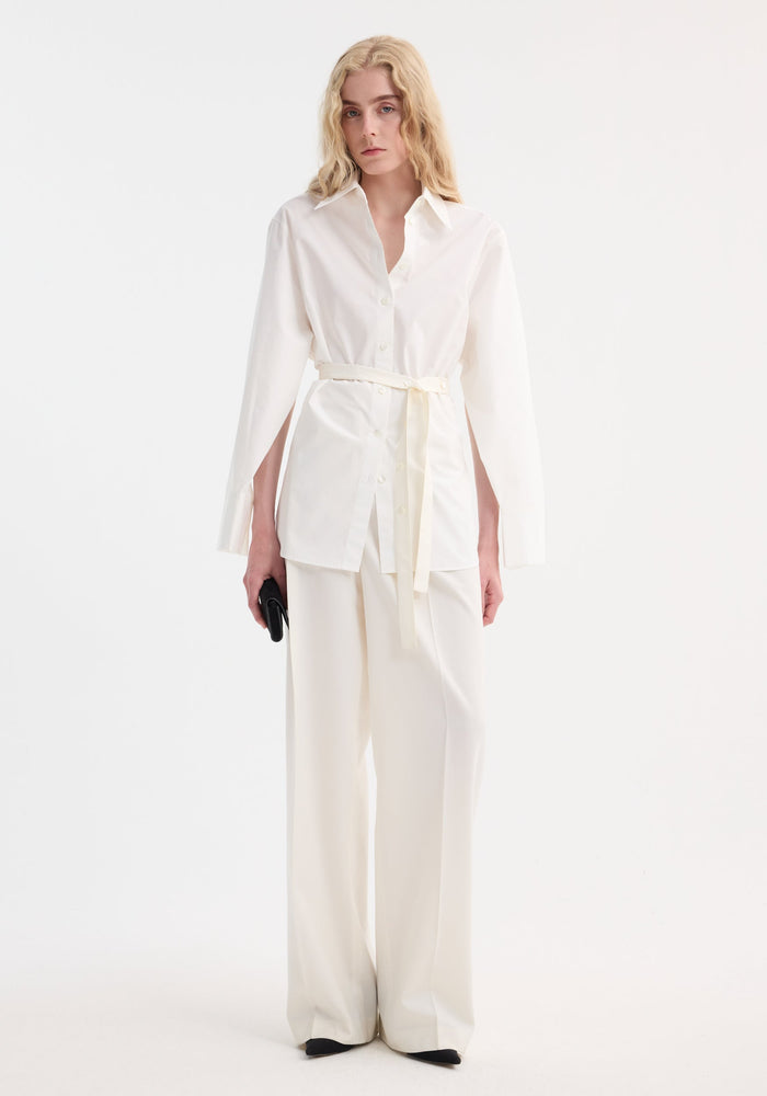 Model in Poplin silk combined gilet-shirt with open side-slit sleeves, wide-leg trousers, black clutch, and shoes; features cotton front, silk back.