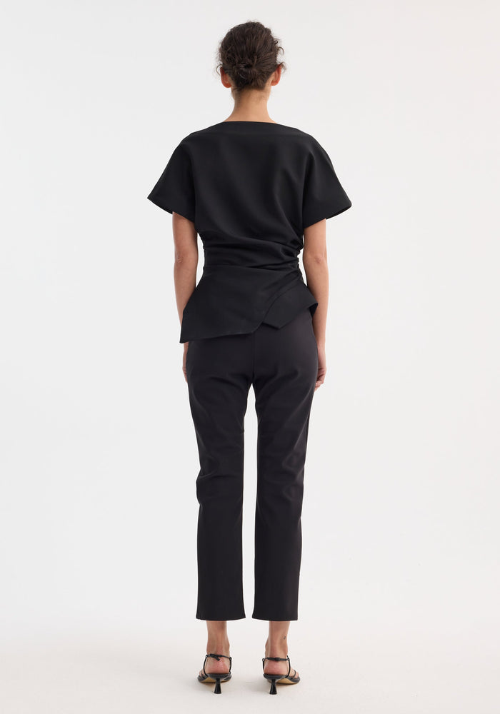 Person wearing a Draped lapel top with sculptural asymmetrical silhouette, slashed neckline, deconstructed lapel, paired with slim trousers and low-heel sandals.