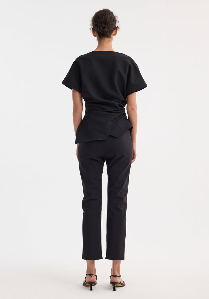 Person wearing a Draped lapel top with sculptural asymmetrical silhouette, slashed neckline, deconstructed lapel, paired with slim trousers and low-heel sandals.