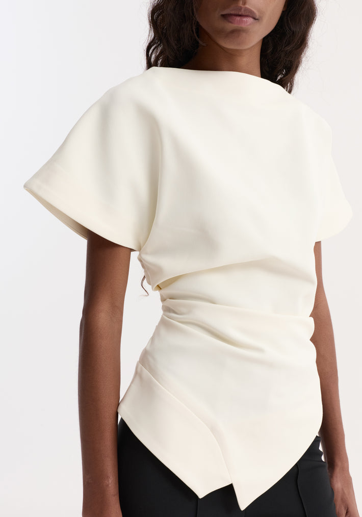 Model wearing Draped lapel top | off white with a slashed neckline, sculpted shoulders, wide sleeves, and an asymmetrical hem, paired with fitted black pants.
