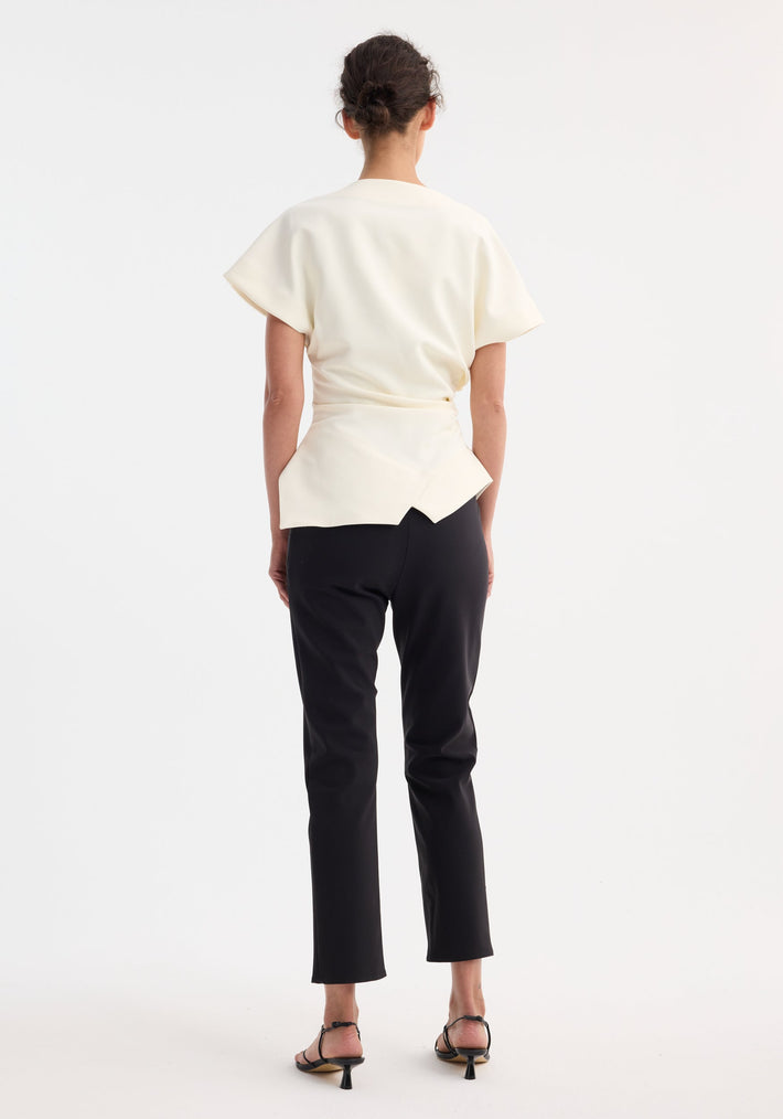 Woman in off-white Draped lapel top with asymmetric hem, sculptural silhouette, worn with black slim-fit trousers and black strappy sandals, hair in a bun.