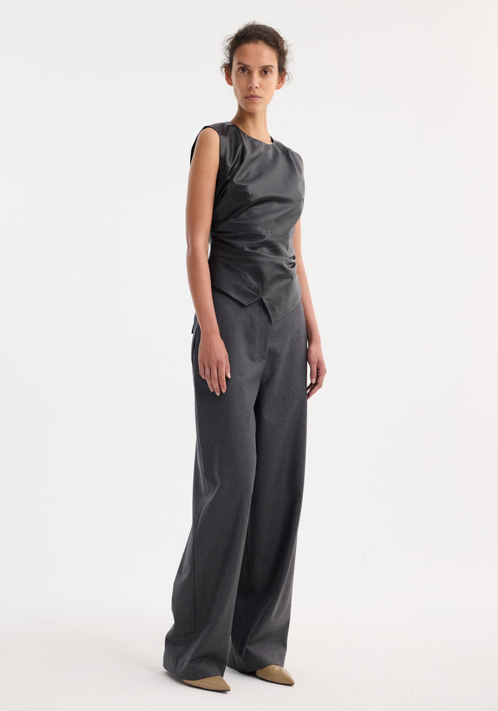 Model wearing Sleeveless draped leather top | black with asymmetrical hem, paired with wide-leg trousers and tan-beige pumps, showcasing modern elegance.