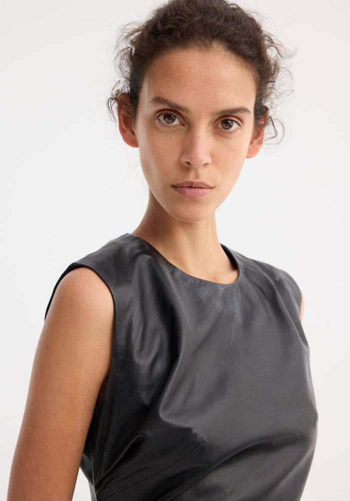 Young woman models the Sleeveless Draped Leather Top in black, featuring an asymmetrical lapel and fitted waist, showcasing modern elegance against a light backdrop.