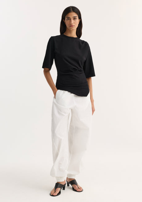 Woman wearing Reinvented twisted t-shirt | black, showcasing its fitted, asymmetrical twist design, paired with loose white trousers and black heeled sandals.