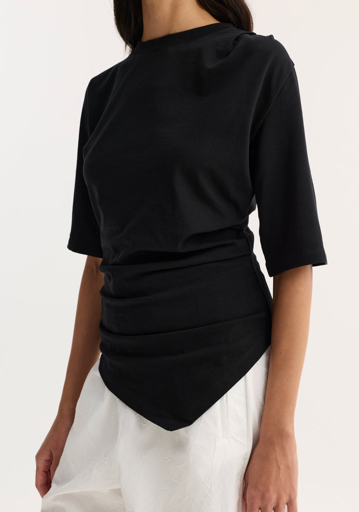 Reinvented twisted t-shirt in black, featuring elbow-length sleeves and a draped, asymmetrical hem, paired with loose, textured white pants against a plain background.