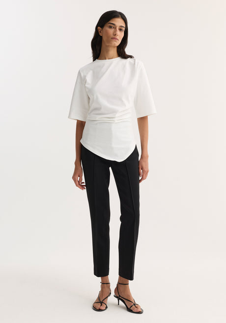 Woman in Reinvented Twisted T-Shirt | Optic White, featuring a sculptural asymmetrical drape and fitted waist, paired with slim-fit trousers and low-heel sandals.