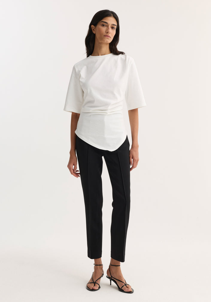 Woman in Reinvented Twisted T-Shirt | Optic White, featuring a sculptural asymmetrical drape and fitted waist, paired with slim-fit trousers and low-heel sandals.