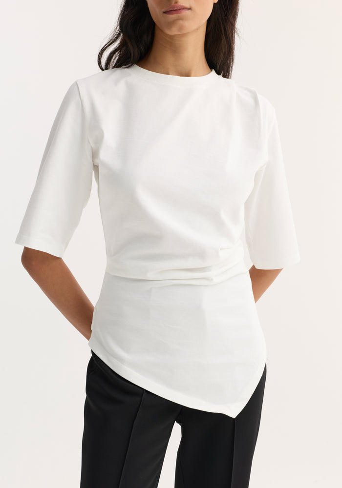 Reinvented twisted t-shirt in optic white, featuring an asymmetrical hem and elbow-length sleeves, worn with black trousers.