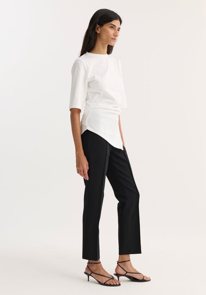 Model wearing Reinvented twisted t-shirt, optic white, with an asymmetrical drape, fitted waist, elbow sleeves, paired with straight-leg trousers and strappy heeled sandals.