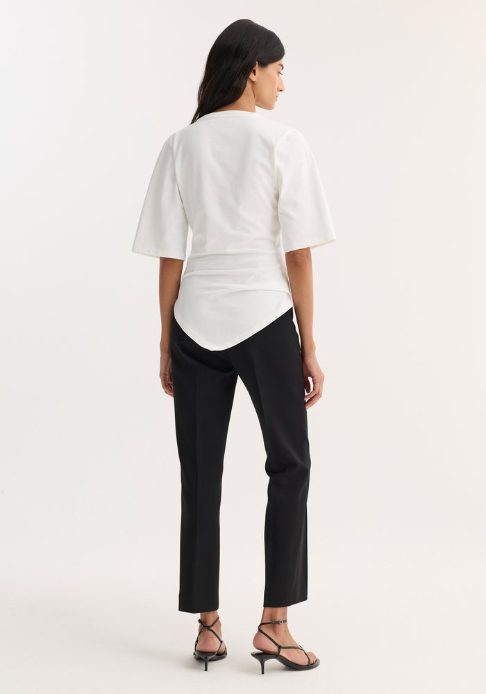 Rear-view of a woman in a Reinvented twisted t-shirt with elbow-length sleeves and pointed hem, paired with slim black trousers and strappy heeled sandals.