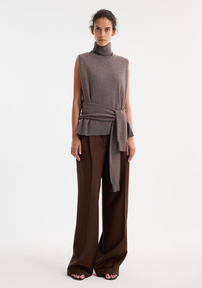 Model wearing Double-layered knit top | mud melange, featuring a sleeveless high-neck with waist tie, and paired with wide-leg chocolate trousers and black sandals.