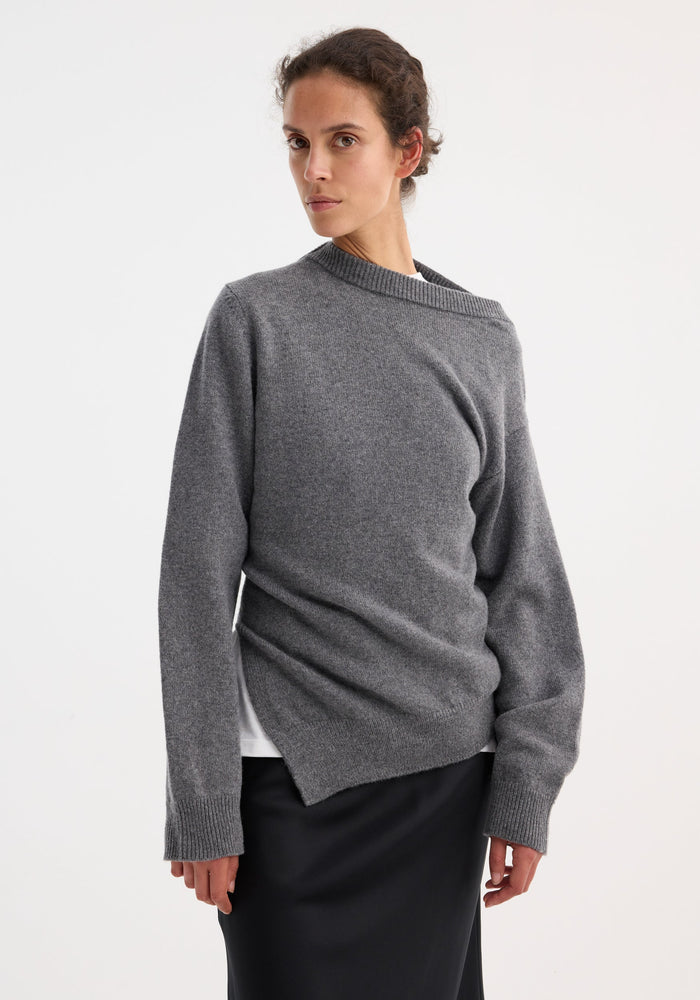 Woman in a mid grey melange two-way deconstructed jumper with an asymmetrical neckline, paired with a black skirt, showcasing minimalist elegance and versatile styling.