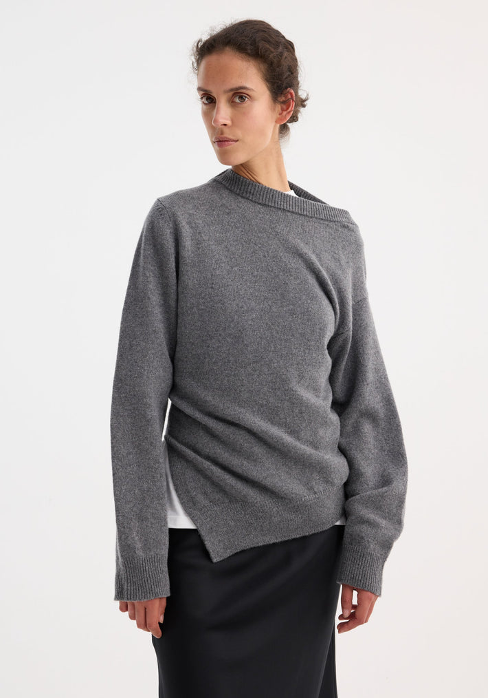 Woman in a mid grey melange two-way deconstructed jumper with an asymmetrical neckline, paired with a black skirt, showcasing minimalist elegance and versatile styling.