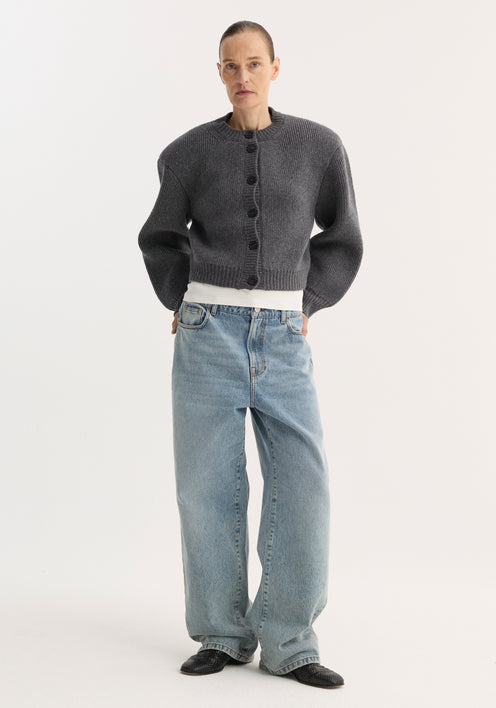 Person models the Statement Shoulder Cardigan in mid grey melange, featuring a structured oversized fit and button closure, paired with wide-leg jeans and pointed shoes.