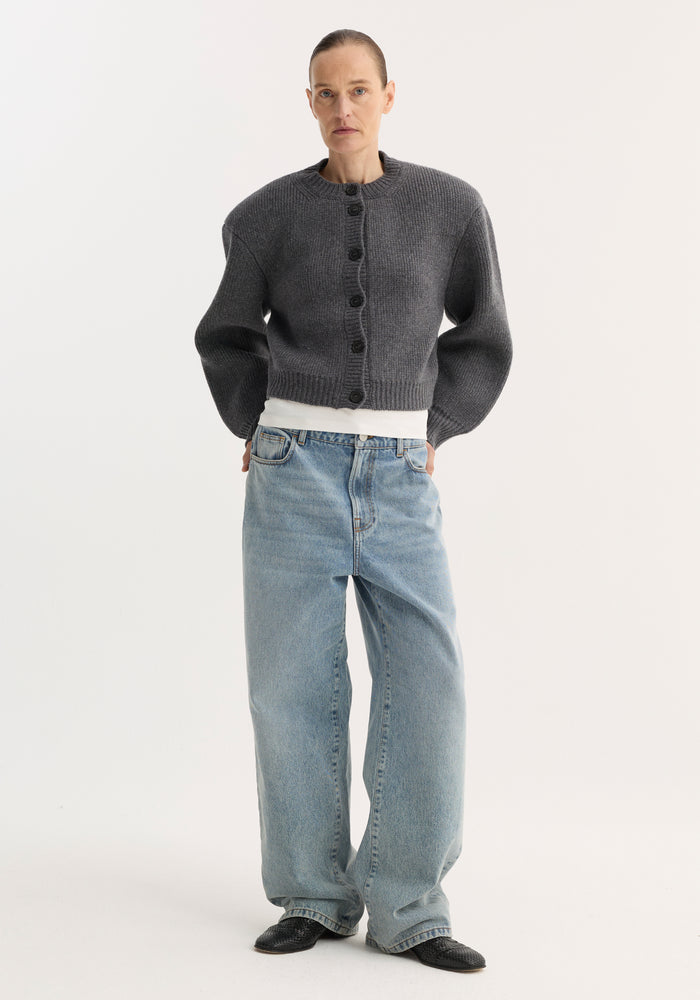 Person models the Statement Shoulder Cardigan in mid grey melange, featuring a structured oversized fit and button closure, paired with wide-leg jeans and pointed shoes.