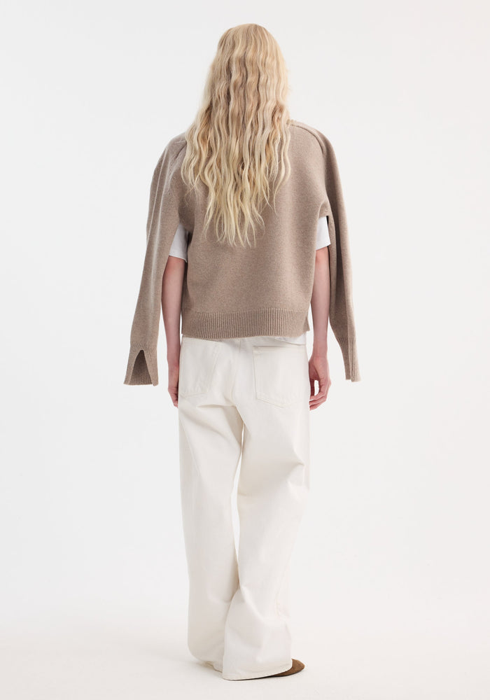Person with long hair models a taupe melange Sleeve wrap jumper, featuring a relaxed fit and wrap-sleeve design, over a white T-shirt and trousers.