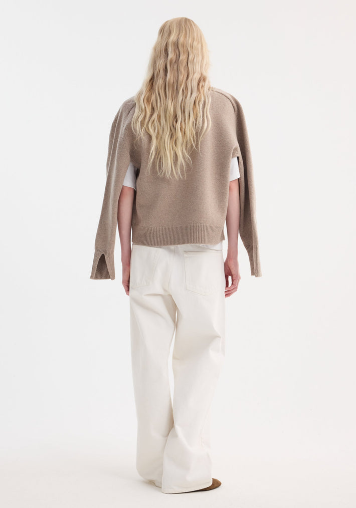 Person with long hair models a taupe melange Sleeve wrap jumper, featuring a relaxed fit and wrap-sleeve design, over a white T-shirt and trousers.