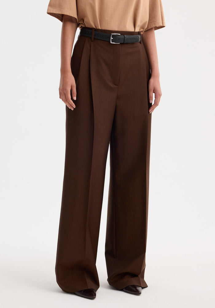 Mid-waist tailored trousers in mahogany with high-waist, wide-leg design, double pleats, and a black leather belt, worn with pointed-toe shoes.