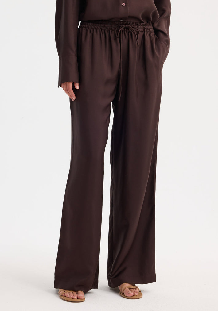 Wide leg silk trousers in mahogany with elastic waistband, drawstring, side pockets, and relaxed fit, made from 100% silk. Model wears with open-toe sandals.